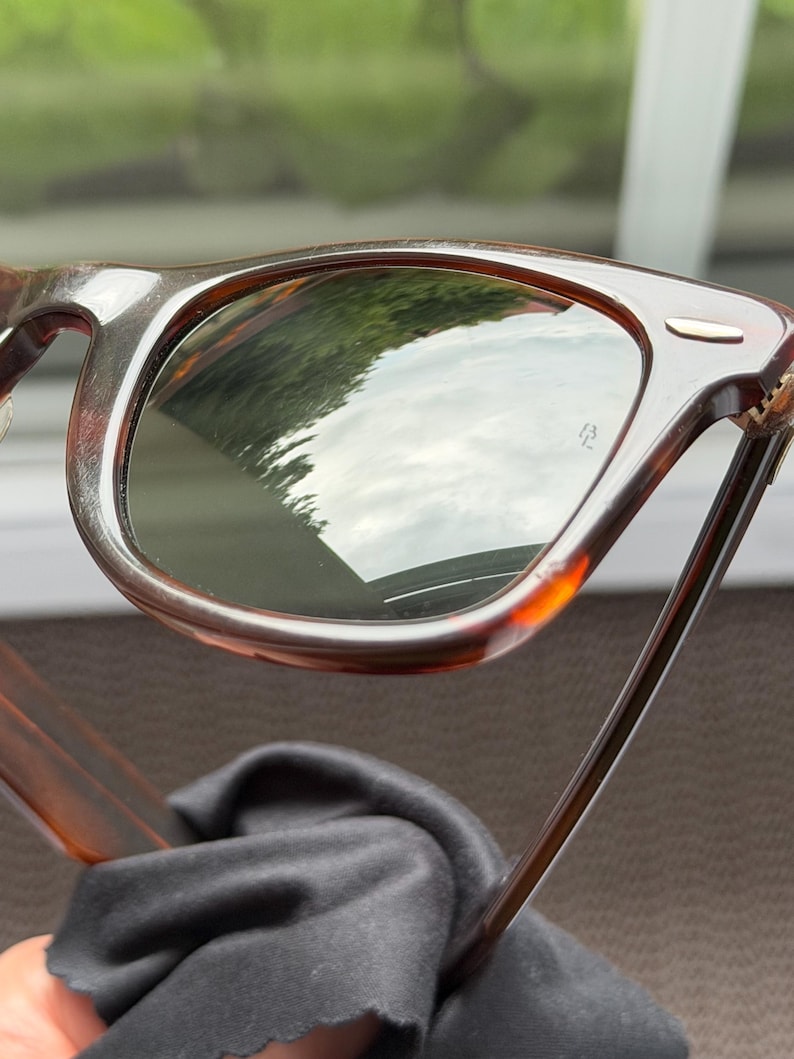 May include: Tortoiseshell-patterned sunglasses with dark green lenses. The frame features a classic rectangular shape with a small silver-toned metal accent on the top corner of the lens rim. The lenses show a reflection of the outdoors.