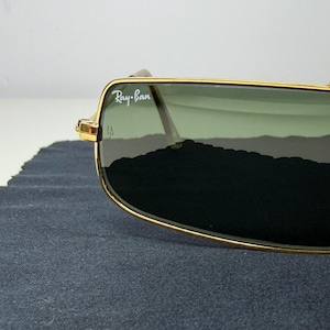 May include: Gold-tone metal frame sunglasses featuring rectangular lenses with a green tint. The upper corner of the lens displays the white Ray-Ban logo. The frame has a thin, polished gold finish with visible hinges and clear nose pads.