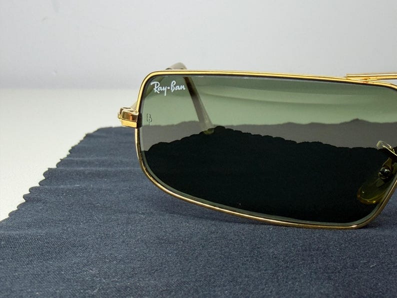 May include: Gold-tone metal frame sunglasses featuring rectangular lenses with a green tint. The upper corner of the lens displays the white Ray-Ban logo. The frame has a thin, polished gold finish with visible hinges and clear nose pads.