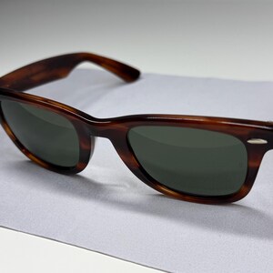 May include: Tortoiseshell-patterned sunglasses with dark green lenses and silver-tone metal accents at the outer corners of the frame. The frames feature a classic wayfarer-style shape with thick rims and matching temples.