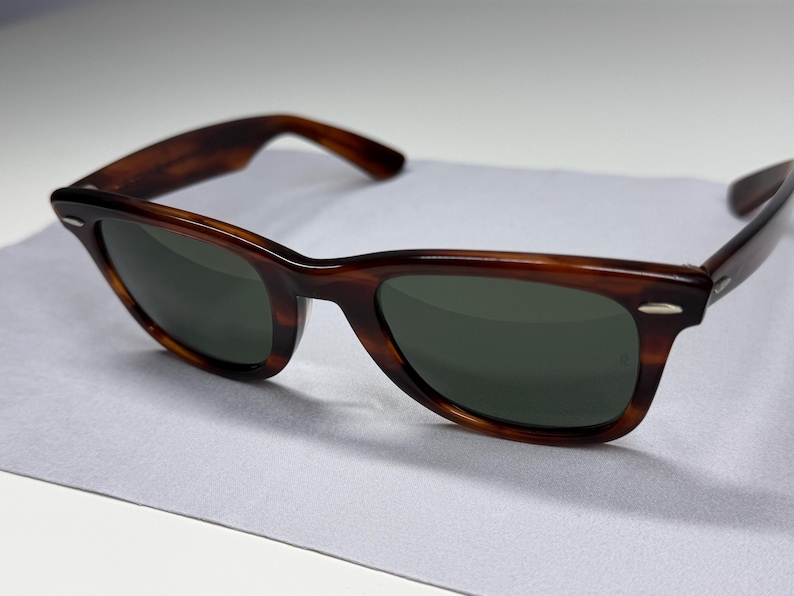 May include: Tortoiseshell-patterned sunglasses with dark green lenses and silver-tone metal accents at the outer corners of the frame. The frames feature a classic wayfarer-style shape with thick rims and matching temples.