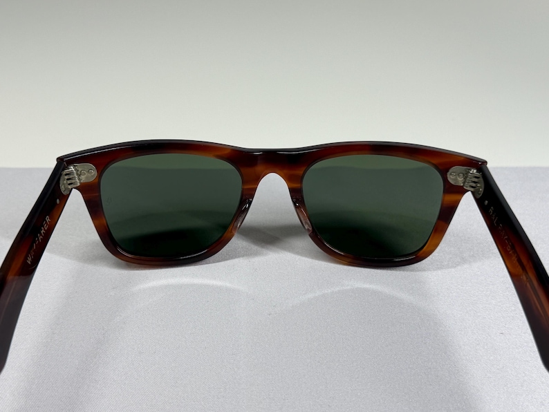May include: Tortoiseshell-patterned sunglasses with dark green lenses. The inner left temple arm features the text Wayfarer, while the inner right temple arm displays B&L 5022. The frame has silver-toned metal hinges connecting the arms to the front.