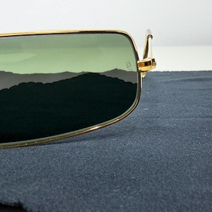 May include: Rectangular sunglasses with a thin gold metal frame and green tinted lenses. The right lens features a small etched logo near the hinge. The frame has a metallic gold finish and the lenses provide a dark green view of the surroundings.
