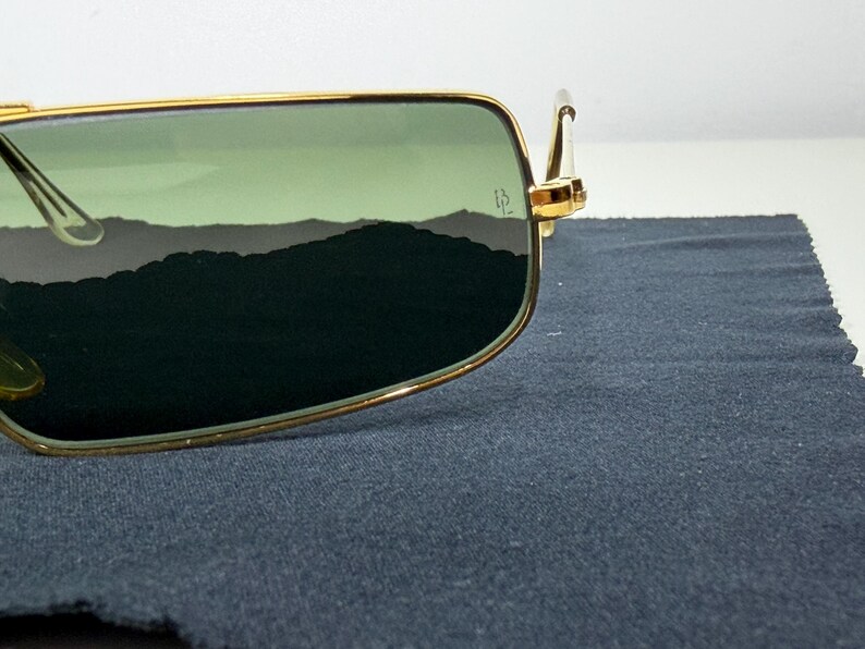May include: Rectangular sunglasses with a thin gold metal frame and green tinted lenses. The right lens features a small etched logo near the hinge. The frame has a metallic gold finish and the lenses provide a dark green view of the surroundings.