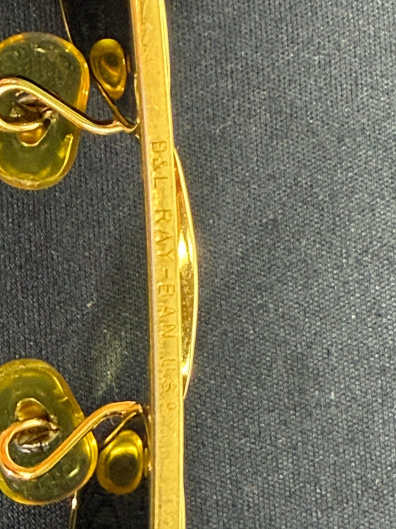 May include: Gold-toned metal eyeglasses frame featuring yellow-tinted nose pads. The bridge is engraved with the text B&L RAY-BAN U.S.A. The frame structure includes thin metal arms and wire components that connect to the nose pads.