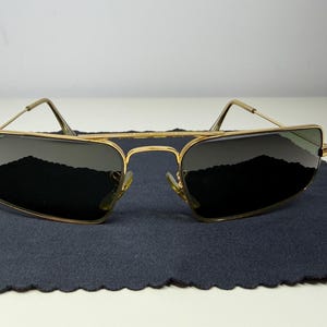 May include: Gold-tone metal frame sunglasses with a rectangular shape and a double bridge design. The lenses are dark tinted, and the Ray-Ban logo is visible on the upper left lens. The thin temples feature curved ends for a secure fit.
