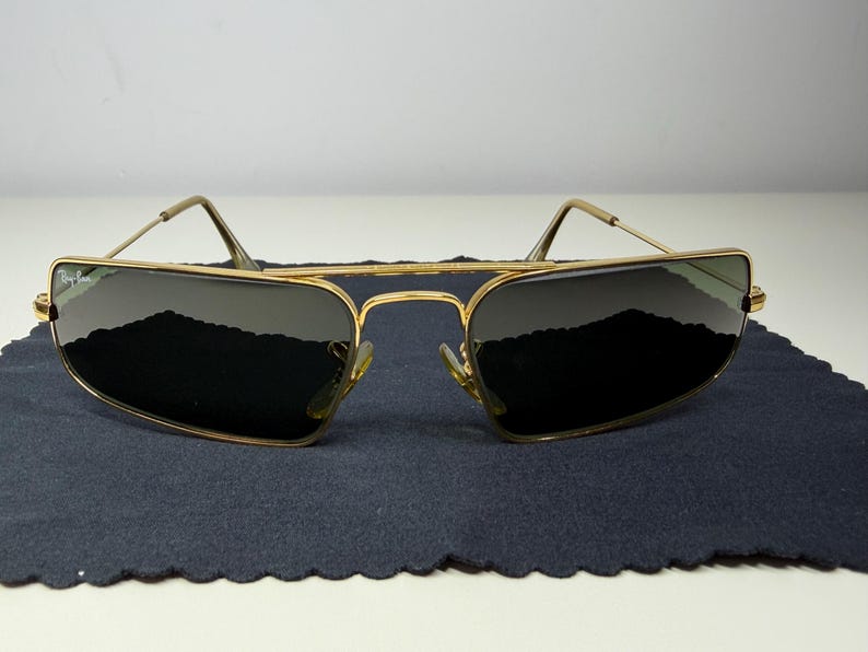 May include: Gold-tone metal frame sunglasses with a rectangular shape and a double bridge design. The lenses are dark tinted, and the Ray-Ban logo is visible on the upper left lens. The thin temples feature curved ends for a secure fit.
