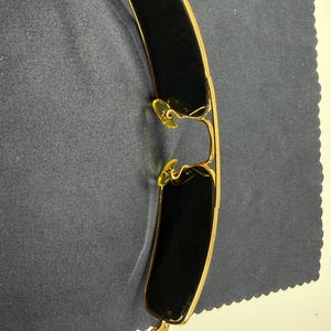 May include: Gold-toned metal sunglasses featuring rectangular dark lenses and a thin wire frame. The eyewear includes adjustable yellow-tinted nose pads and slender temples, showcasing a classic construction with a metallic finish and dark lenses.
