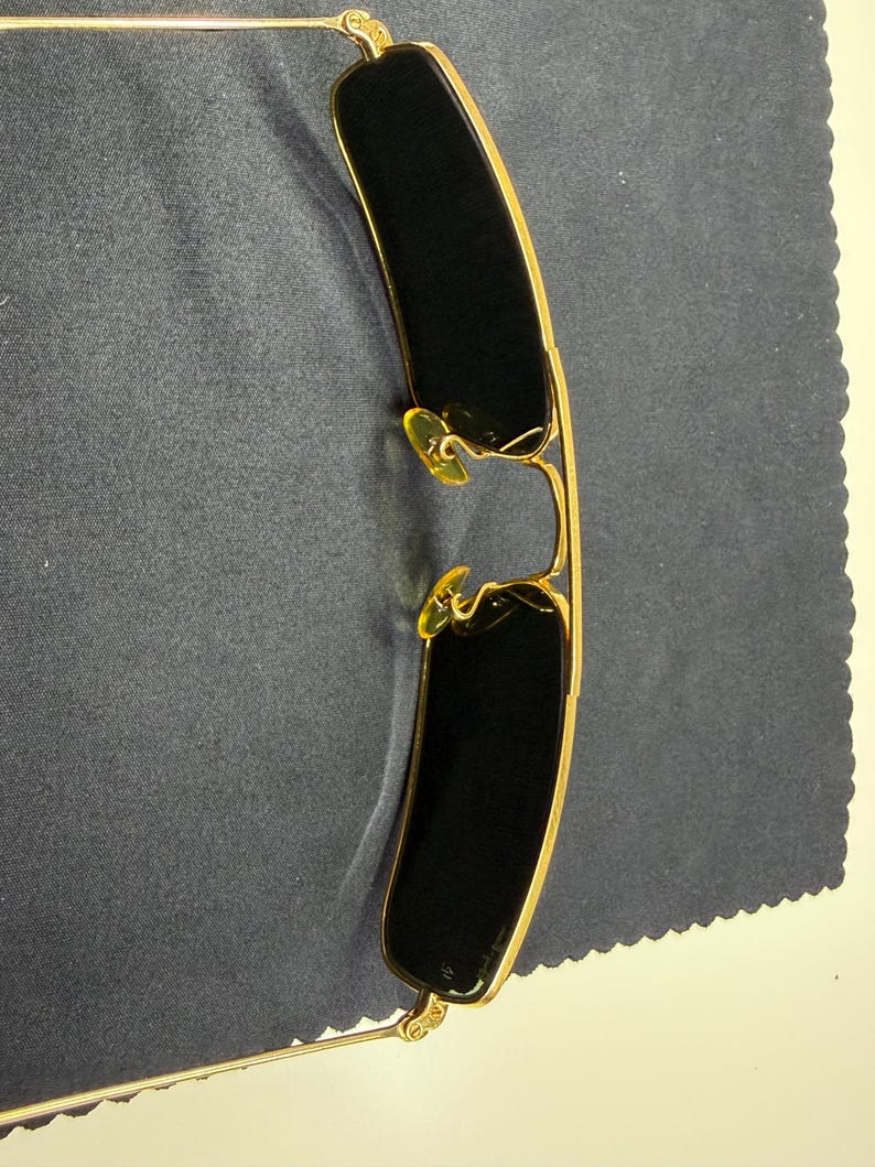 May include: Gold-toned metal sunglasses featuring rectangular dark lenses and a thin wire frame. The eyewear includes adjustable yellow-tinted nose pads and slender temples, showcasing a classic construction with a metallic finish and dark lenses.