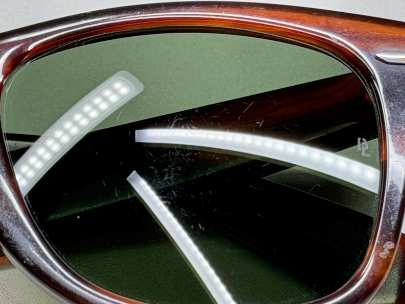 May include: Tortoiseshell-patterned sunglasses featuring dark green lenses. The frame has a polished finish with a visible silver-tone metal accent at the temple hinge. The lenses show light surface scratches and reflections of light strips.