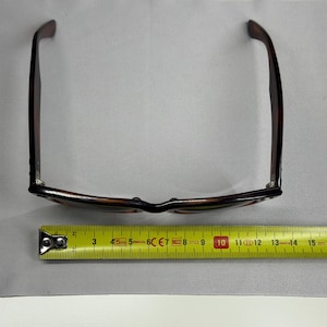 May include: Tortoiseshell-patterned sunglasses with dark brown frames and temples, positioned above a yellow measuring tape that indicates a width of approximately 5.5 inches across the front of the frame from hinge to hinge.