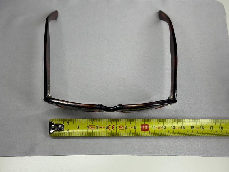 May include: Tortoiseshell-patterned sunglasses with dark brown frames and temples, positioned above a yellow measuring tape that indicates a width of approximately 5.5 inches across the front of the frame from hinge to hinge.