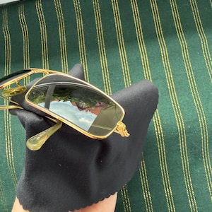 May include: Rectangular sunglasses with a thin gold-tone metal frame and dark tinted lenses. The sunglasses are resting on a black microfiber cleaning cloth. The lenses show a reflection of an outdoor scene with a building and a blue sky.