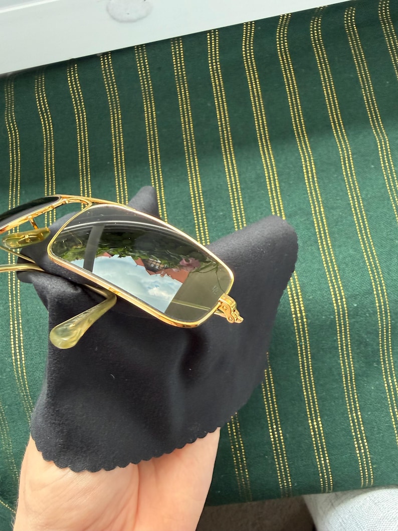 May include: Rectangular sunglasses with a thin gold-tone metal frame and dark tinted lenses. The sunglasses are resting on a black microfiber cleaning cloth. The lenses show a reflection of an outdoor scene with a building and a blue sky.
