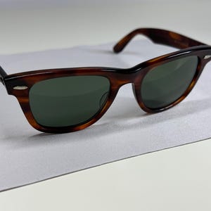 May include: Tortoiseshell sunglasses featuring a classic wayfarer-style frame with dark green lenses. The frame has a polished brown and black mottled pattern with silver-tone metal accents at the hinges on both sides of the front frame.