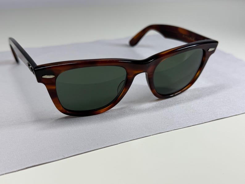 May include: Tortoiseshell sunglasses featuring a classic wayfarer-style frame with dark green lenses. The frame has a polished brown and black mottled pattern with silver-tone metal accents at the hinges on both sides of the front frame.