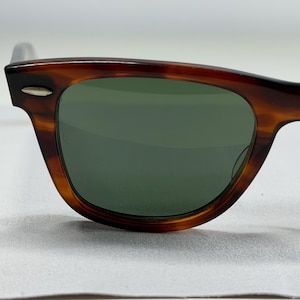 May include: Tortoiseshell-patterned sunglasses with dark green lenses. The frames feature a classic rectangular shape with a distinct brown and black mottled finish. A small, silver-toned metal accent is visible on the upper corner of the frame.