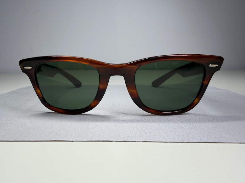 May include: Sunglasses with a brown tortoiseshell frame and dark green lenses. The frame features a classic wayfarer shape with silver-toned metal accents on the outer corners of the front rims, providing a clean and functional eyewear design.