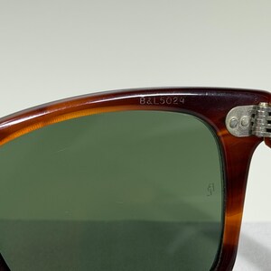 May include: Tortoiseshell-patterned sunglasses with dark green lenses. The upper inner frame is engraved with the text B&L 5024. A silver-toned metal hinge with three rivets connects the frame to the temple. A small logo is etched on the lens.