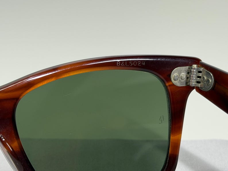 May include: Tortoiseshell-patterned sunglasses with dark green lenses. The upper inner frame is engraved with the text B&L 5024. A silver-toned metal hinge with three rivets connects the frame to the temple. A small logo is etched on the lens.