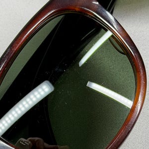 May include: Sunglasses with a dark brown tortoise shell pattern frame and dark green tinted lenses. The frame has a rounded rectangular shape with a smooth, polished finish. The lenses show reflections of light sources, highlighting the surface.