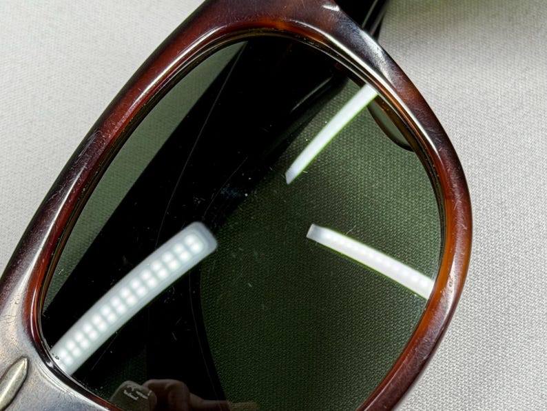 May include: Sunglasses with a dark brown tortoise shell pattern frame and dark green tinted lenses. The frame has a rounded rectangular shape with a smooth, polished finish. The lenses show reflections of light sources, highlighting the surface.