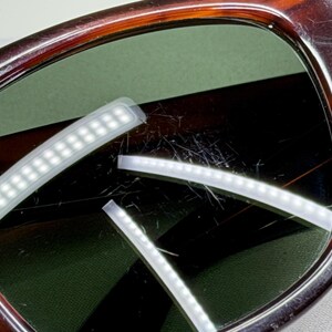 May include: Tortoiseshell-patterned eyeglasses featuring dark brown and amber frames with green-tinted lenses. The lenses show visible surface scratches and reflections from rectangular light sources, with a small logo etched on the left lens.