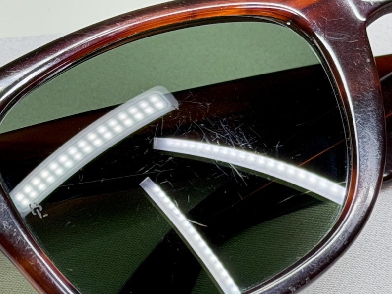 May include: Tortoiseshell-patterned eyeglasses featuring dark brown and amber frames with green-tinted lenses. The lenses show visible surface scratches and reflections from rectangular light sources, with a small logo etched on the left lens.