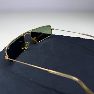 May include: Gold-tone metal frame sunglasses featuring rectangular lenses with a dark green tint. The frames include a thin double bridge design and slender temples with clear plastic tips. The Ray-Ban logo is visible on the upper right lens.