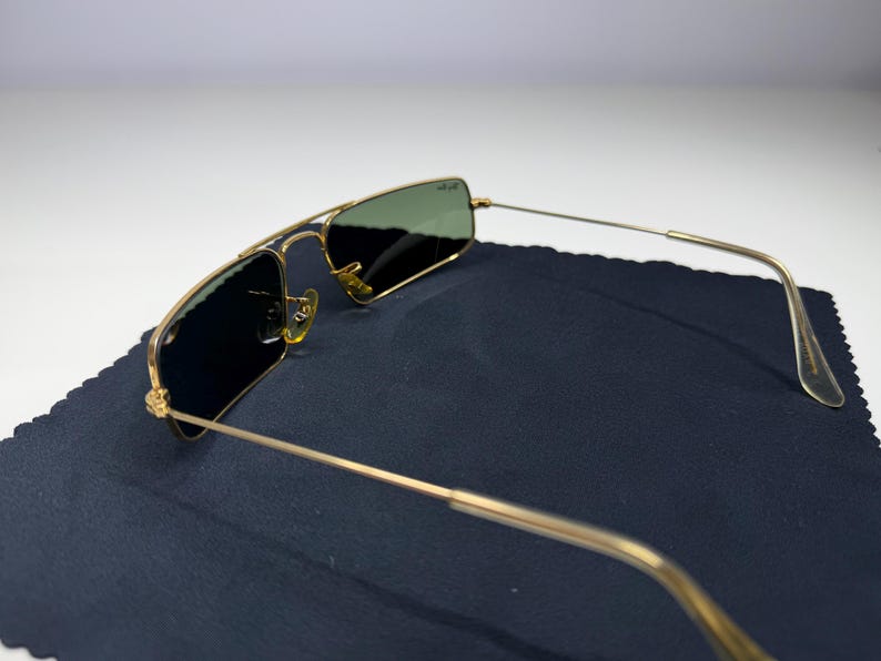 May include: Gold-tone metal frame sunglasses featuring rectangular lenses with a dark green tint. The frames include a thin double bridge design and slender temples with clear plastic tips. The Ray-Ban logo is visible on the upper right lens.