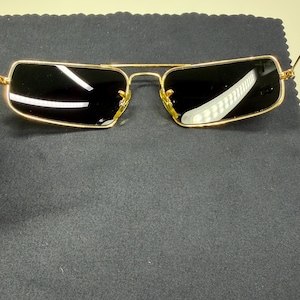 May include: Rectangular sunglasses featuring a thin gold-tone metal frame and dark tinted lenses. The bridge connects the two lenses, and the temples extend outward. The Ray-Ban logo is visible on the upper corner of the right lens.