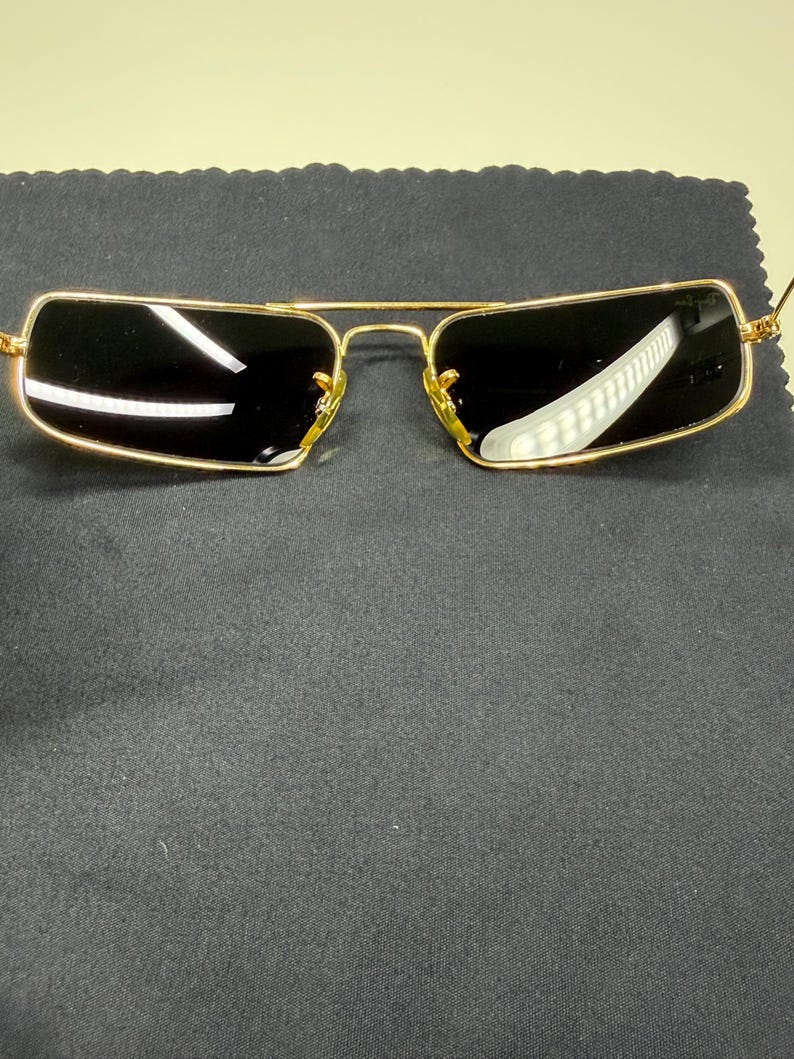 May include: Rectangular sunglasses featuring a thin gold-tone metal frame and dark tinted lenses. The bridge connects the two lenses, and the temples extend outward. The Ray-Ban logo is visible on the upper corner of the right lens.