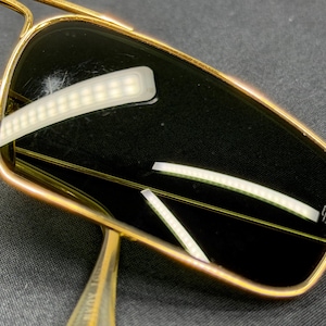 May include: Rectangular sunglasses featuring a polished gold-tone metal frame and dark tinted lenses. The right lens displays a small white logo mark in the corner. The thin metal temples extend from the frame, showcasing a metallic finish.