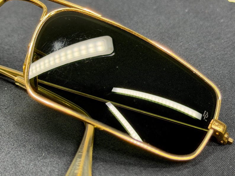 May include: Rectangular sunglasses featuring a polished gold-tone metal frame and dark tinted lenses. The right lens displays a small white logo mark in the corner. The thin metal temples extend from the frame, showcasing a metallic finish.