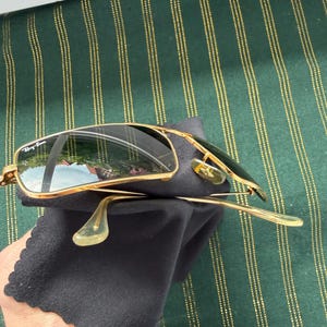 May include: Gold-tone metal frame sunglasses with dark, reflective lenses. The left lens features the white Ray-Ban logo in the upper corner. The sunglasses have thin gold temples with clear plastic tips and adjustable nose pads, resting on a black cloth.