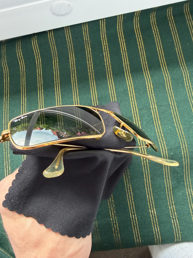 May include: Gold-tone metal frame sunglasses with dark, reflective lenses. The left lens features the white Ray-Ban logo in the upper corner. The sunglasses have thin gold temples with clear plastic tips and adjustable nose pads, resting on a black cloth.