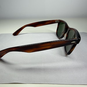 May include: Tortoiseshell sunglasses featuring a classic frame design with dark brown and amber mottled patterns. The eyewear includes dark green tinted lenses and silver-toned metal accents on the outer corners of the frame front.