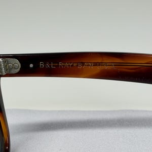 May include: Tortoiseshell-patterned sunglasses featuring a brown frame with a metallic hinge. The inner temple arm is clearly engraved with the text B&L RAY-BAN U.S.A. in a light-colored font, indicating the manufacturer and origin of the eyewear.