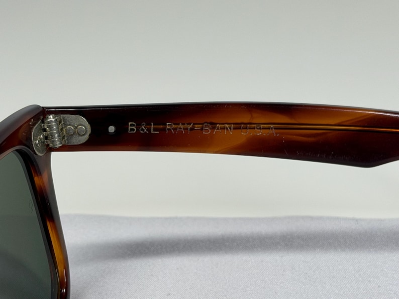May include: Tortoiseshell-patterned sunglasses featuring a brown frame with a metallic hinge. The inner temple arm is clearly engraved with the text B&L RAY-BAN U.S.A. in a light-colored font, indicating the manufacturer and origin of the eyewear.