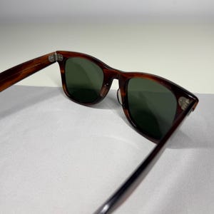 May include: Tortoiseshell-patterned sunglasses with dark green lenses. The frames feature a classic rectangular shape with rounded edges and silver-toned metal rivets on the front corners. The temples are connected to the frame with metal hinges.