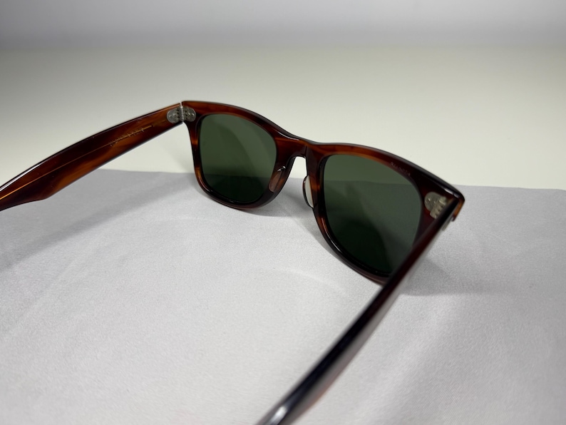 May include: Tortoiseshell-patterned sunglasses with dark green lenses. The frames feature a classic rectangular shape with rounded edges and silver-toned metal rivets on the front corners. The temples are connected to the frame with metal hinges.