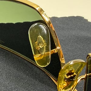 May include: Gold-tone metal sunglasses featuring green-tinted lenses and clear, elongated nose pads. The inner lens surface displays a subtle logo detail, while the frame construction highlights a metallic finish and visible hinge components.