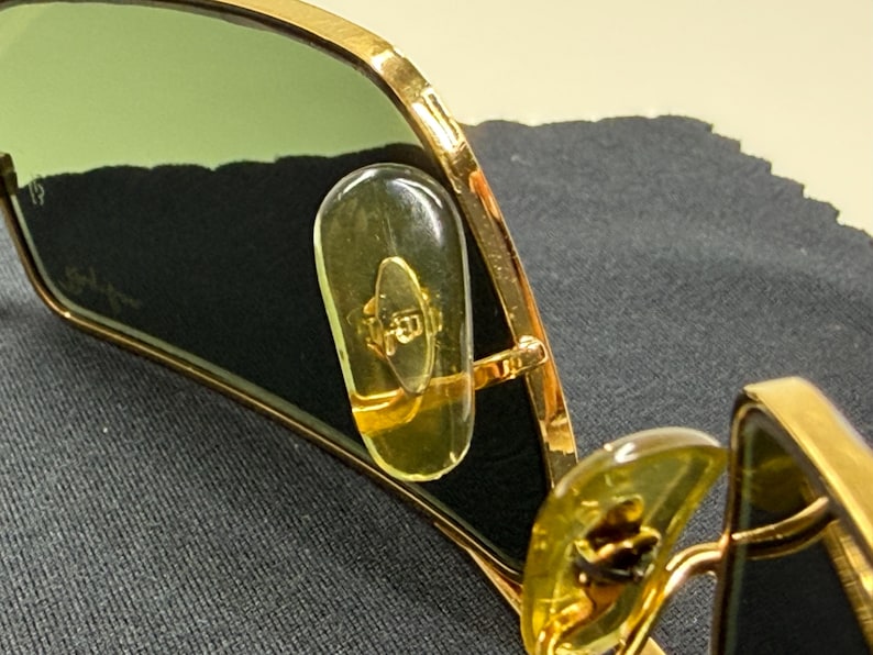 May include: Gold-tone metal sunglasses featuring green-tinted lenses and clear, elongated nose pads. The inner lens surface displays a subtle logo detail, while the frame construction highlights a metallic finish and visible hinge components.