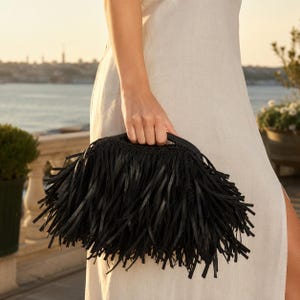 May include: A black clutch bag featuring a dense layer of long, thin, dangling fringe strips covering the entire exterior surface. The bag has a curved top edge with a structured handle integrated into the design, creating a textured appearance.