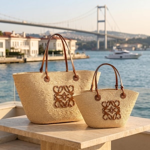 May include: Two woven straw tote bags in different sizes, each featuring tan leather handles and a prominent brown leather logo patch on the front. The bags have a natural light beige texture and are positioned on a light-colored stone surface.