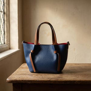 K&ouml;nnte beinhalten: Eine marineblaue Handtasche aus genarbtem Leder mit hellbraunen Ledergriffen und passenden seitlichen Knotendetails. Das Innenfutter ist leuchtend rot und die Tasche verf&uuml;gt &uuml;ber silberfarbene Beschl&auml;ge zur Befestigung eines Schulterriemens.