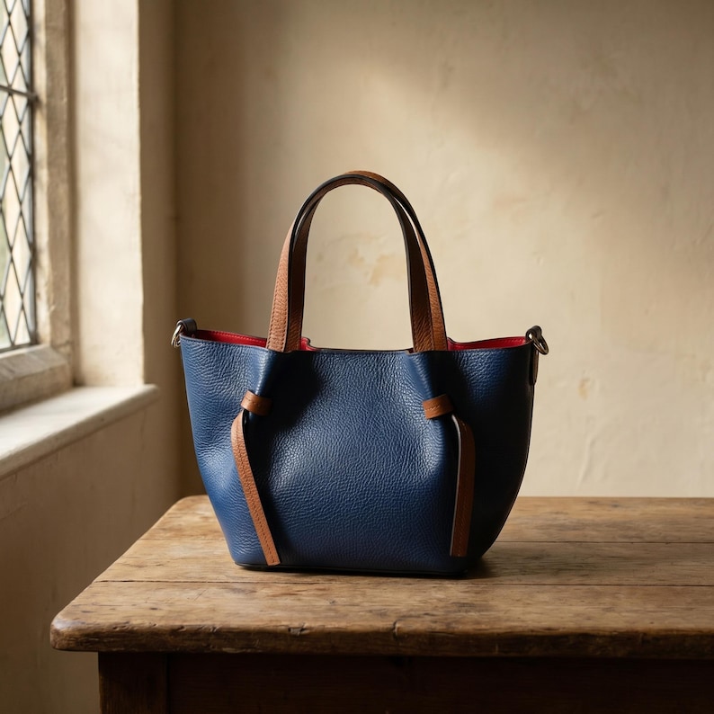 K&ouml;nnte beinhalten: Eine marineblaue Handtasche aus genarbtem Leder mit hellbraunen Ledergriffen und passenden seitlichen Knotendetails. Das Innenfutter ist leuchtend rot und die Tasche verf&uuml;gt &uuml;ber silberfarbene Beschl&auml;ge zur Befestigung eines Schulterriemens.