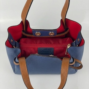 May include: A blue handbag with a textured surface, featuring tan leather handles and a vibrant red interior lining. The bag includes a central zippered compartment, gold-toned metal snap closures, and a structured open-top design with side gussets.