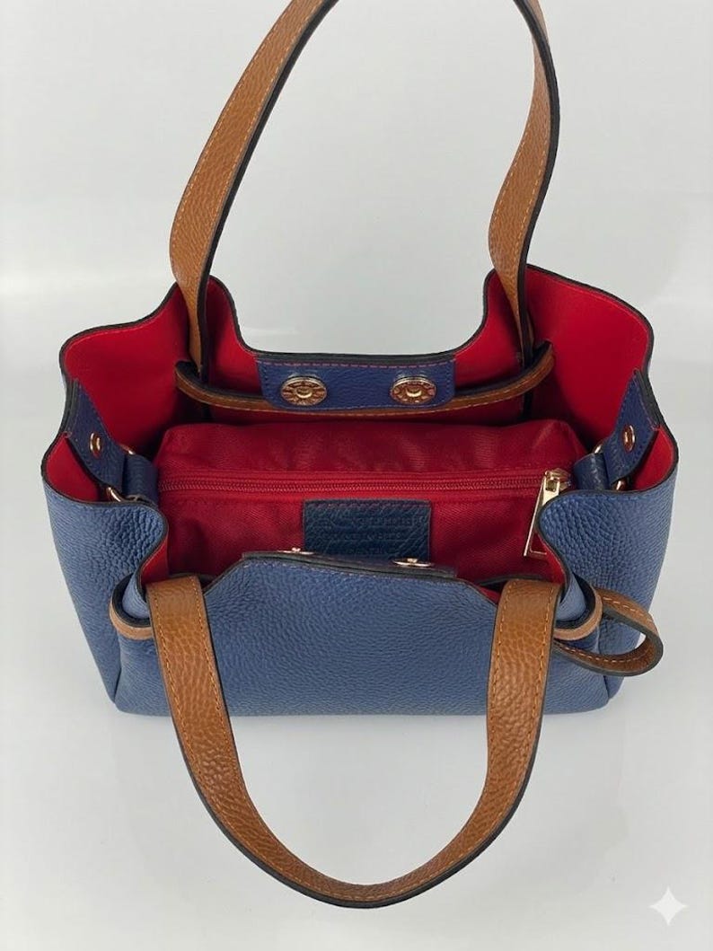 May include: A blue handbag with a textured surface, featuring tan leather handles and a vibrant red interior lining. The bag includes a central zippered compartment, gold-toned metal snap closures, and a structured open-top design with side gussets.