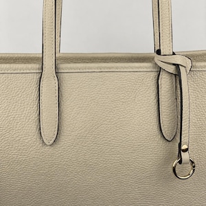 Op de afbeelding: Een beige tote bag met een generfde textuur, voorzien van twee verticale schouderbanden bevestigd aan de bovenrand. Een dun leren koord is rond &eacute;&eacute;n band geknoopt en eindigt in een kleine goudkleurige metalen ring met bijpassende klinknagel.